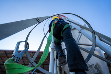 A worker or engineer, wearing essential safety equipment including a harness and a yellow hard hat, is captured climbing a fixed vertical ladder on the side of a large industrial silo.
