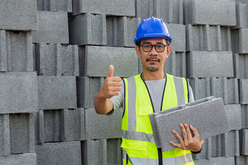 A cheerful Asian construction worker stands in front of a large pile of concrete blocks. He holds one block and gives a confident thumbs up. Quality control, acceptance at construction site.