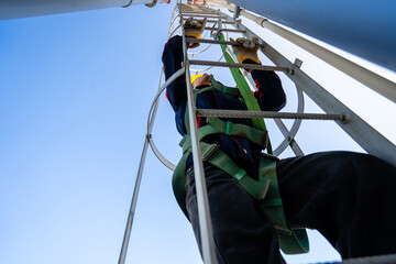 A worker or engineer, wearing essential safety equipment including a harness and a yellow hard hat, is captured climbing a fixed vertical ladder on the side of a large industrial silo.