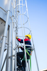 A worker or engineer, wearing essential safety equipment including a harness and a yellow hard hat, is captured climbing a fixed vertical ladder on the side of a large industrial silo.