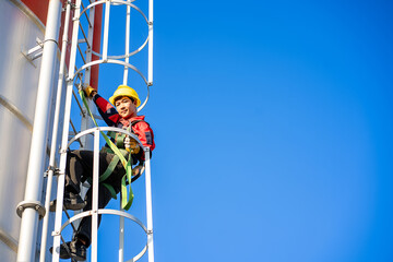 A worker or engineer, wearing essential safety equipment including a harness and a yellow hard hat, is captured climbing a fixed vertical ladder on the side of a large industrial silo.