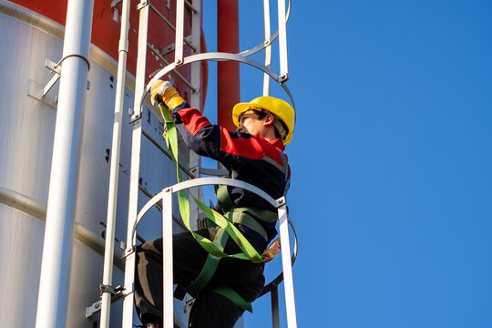 A worker or engineer, wearing essential safety equipment including a harness and a yellow hard hat, is captured climbing a fixed vertical ladder on the side of a large industrial silo.