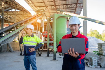 Two construction or factory engineers, wearing protective gear, stand inside an industrial processing plant. One is focused on a laptop, while the other communicates via a walkie-talkie.