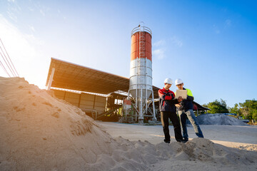 Two construction plant engineers wearing protective gear, stand inside an industrial processing plant. One is focused on a laptop the other communicates via walkie-talkie. digital project management.