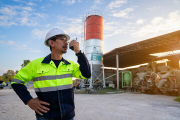 An Asian construction site engineer, wearing protective gear, communicates via walkie-talkie in a construction material yard, with a large cement silo and a concrete mixer truck in the background.