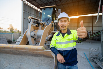 A Professional Asian site manager stands confidently with hands on hips in front of a large yellow wheel loader or heavy machinery in a material handling shed.