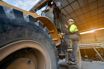 Asian male engineer wearing protective gear climbs into the passenger compartment of a large yellow wheel loader. Heavy machinery and workers working inside a material storage shed.