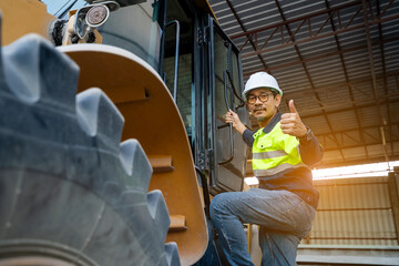 A Professional Asian site manager stands confidently with hands on hips in front of a large yellow wheel loader or heavy machinery in a material handling shed.