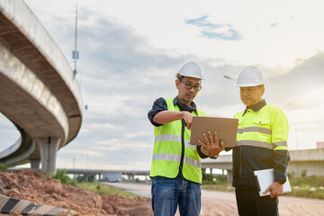 Two construction engineers, wearing hard hats and high-visibility vests, stand at a highway interchange construction site. discussing plans with his colleague who holds a walkie-talkie and tablet.
