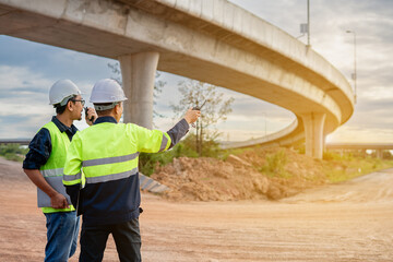 Two construction engineers, wearing hard hats and high-visibility vests, stand at a highway interchange construction site. discussing plans with his colleague who holds a walkie-talkie and tablet.
