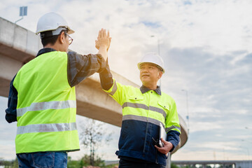 A close-up of two construction or business professionals in hard hats and reflective safety vests shaking hands at an outdoor site, with a bridge structure in the soft-focus background.
