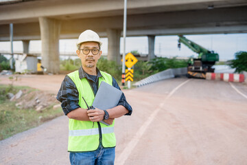A professional Asian construction engineer, stands confidently on a dirt road at an infrastructure site. holds a laptop and a walkie-talkie. The highway bridge structure and heavy machinery