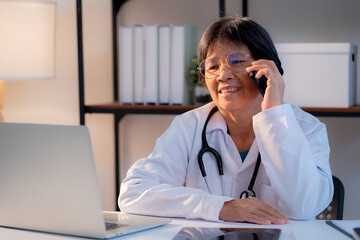 Senior asian doctor speak on smartphone while working on laptop in medical office, offering telemedicine consultation, healthcare and digital technology, elderly female physician online consultation.