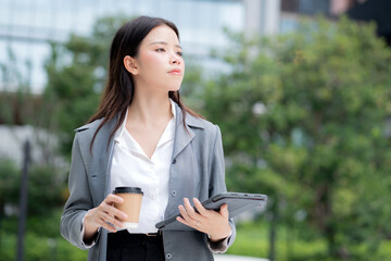 Happy young asian businesswoman standing holding coffee while using tablet outdoors city, digital nomad, business woman or manager touching tablet for working, modern technology, digital device.
