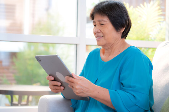 Senior asian woman relax on sofa reading tablet in living room at home, online connection and digital lifestyle, elder watching tablet for social media, remote learning, elderly using chatbot or AI. - Powered by Adobe