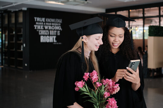 Two diverse female graduate students, teenage friends watching celebration photos on mobile phone together, degree achievement milestones, happiness in academic college, and cheerful congratulations.