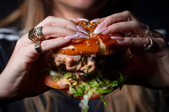 Close-up shot of a woman's hand holding a fat, juicy hamburger on a dark background 