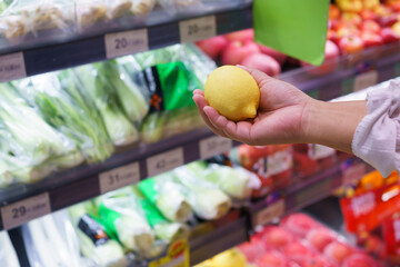 Customer holding fresh lemon while shopping in a grocery store with colorful fruit shelves, healthy lifestyle, clean eating, and fresh food concept.