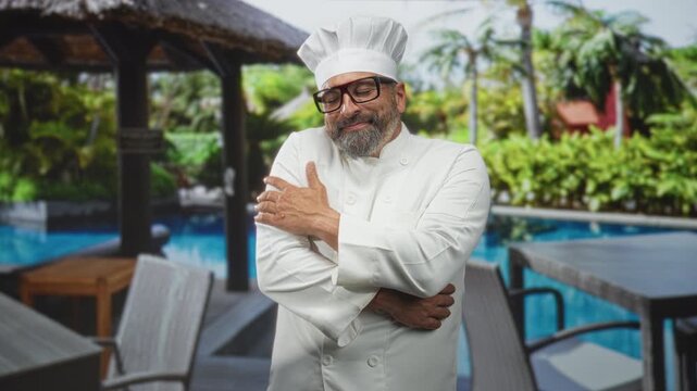 Man chef in white uniform hugs his shoulder with both hands at a poolside dining area near a thatch gazebo; quiet contentment.