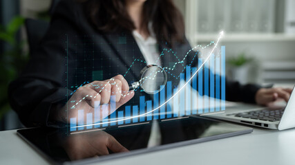 A business professional uses a magnifying glass to examine financial data trends displayed on a digital tablet, showcasing analysis and strategic insights in a modern office environment. Impute