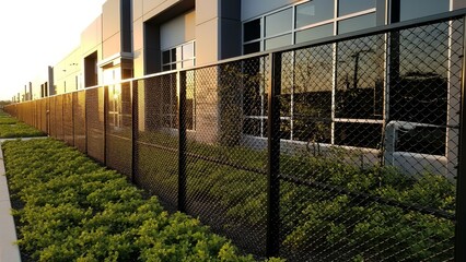 A long black metal security fence with green landscaping alongside a modern industrial building under a warm sunset sky.