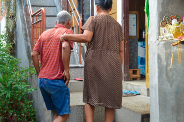 Back-view of an elderly couple in a courtyard, with the wife in a brown polka-dot dress placing her hand on her husband's arm as they walk up concrete steps.