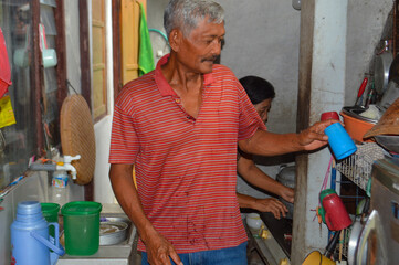 A busy kitchen scene where the elderly man places a clean blue plastic cup onto a shelf, while his wife is partially visible in the background attending to a stove.