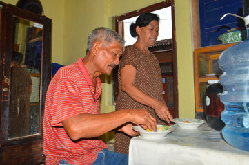 An elderly couple stands and sits near a simple table, with the man preparing his plate and the woman serving rice and side dishes for their morning meal.