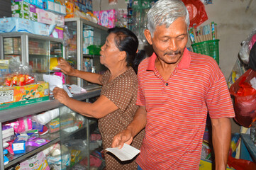 A busy scene in a small home shop where the elderly man holds a receipt while his wife checks items behind a cluttered glass shelf to confirm their stock.