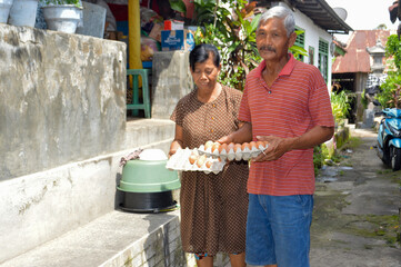An elderly couple stands side-by-side in their outdoor yard, both holding trays of brown chicken eggs they collected from their poultry coops earlier in the day.