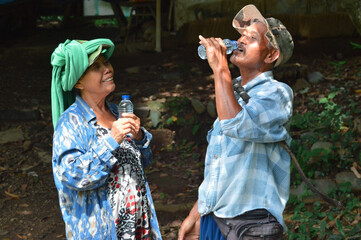 An elderly man in a plaid shirt and cap smiles while drinking from a plastic water bottle, held high above his head, as his wife stands beside him holding her own bottle and smiling.