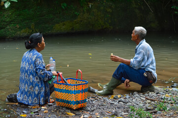 An elderly man in a plaid shirt and boots sits on a rocky riverbank eating a snack, while his wife sits facing him with a water bottle, enjoying a peaceful break in the green environment.