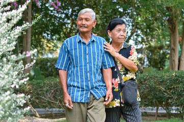 Fototapeta premium A mid-shot portrait of an elderly couple standing together in a park, surrounded by decorative shrubs and pink flowers, the woman gently places her hand on the manâ€™s shoulder as they look forward.