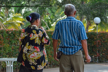 A rear view of an elderly couple walking down a paved path in a garden, the man wears a striped shirt and khaki pants, while the woman in a floral shirt walks beside him toward greenery.