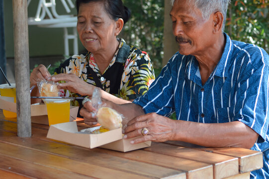 A close-up of an elderly couple sitting at an outdoor wooden cafe table in a park, unwrapping and eating snacks from cardboard boxes, with yellow drinks in plastic cups nearby.