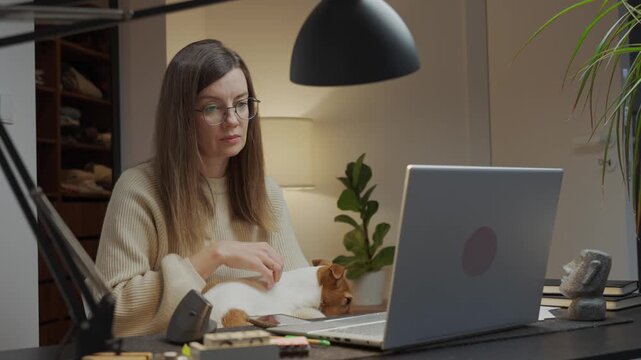 Woman sitting at home desk, working on laptop at home office with small dog on her lap. Person using computer at room with jack russell terrier. Life with pet concept