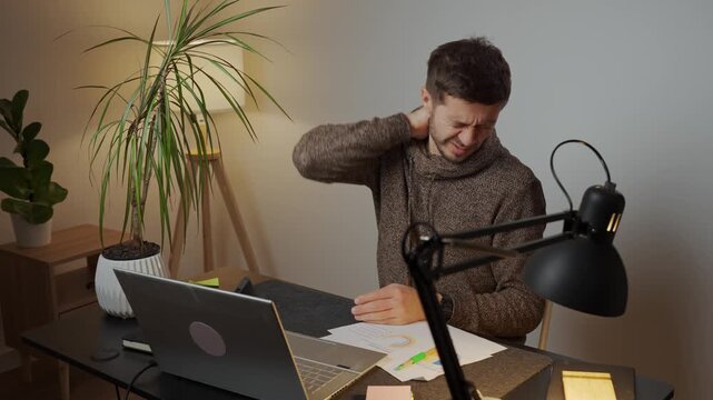 Man sitting at a home desk holding his neck, suffering from pain while working on a laptop. Concept of ergonomic posture and discomfort due to working on computer