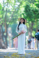 Obraz premium A young Vietnamese woman wearing a traditional white ao dai and a conical hat smiles gracefully under bright sunlight, on street in Hanoi, Vietnam.