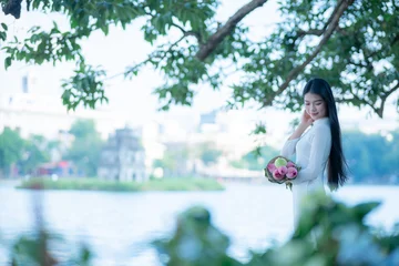 Fotobehang Lotusbloem A young Vietnamese woman in a white ao dai standing by Hoan Kiem Lake, holding a bouquet of pink lotus flowers  © CravenA