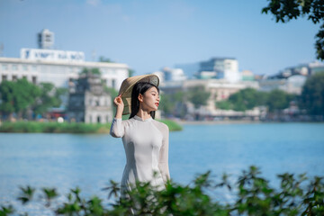 A young Vietnamese woman in a white ao dai and a conical hat standing by Hoan Kiem Lake in Hanoi, Vietnam