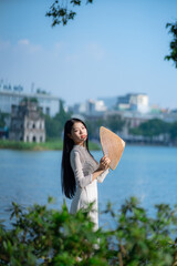 A young Vietnamese woman in a white ao dai and a conical hat standing by Hoan Kiem Lake in Hanoi, Vietnam