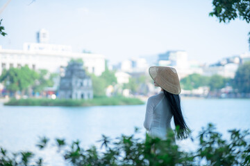 A young Vietnamese woman in a white ao dai and a conical hat standing by Hoan Kiem Lake in Hanoi, Vietnam