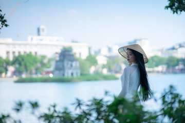 A young Vietnamese woman in a white ao dai and a conical hat standing by Hoan Kiem Lake in Hanoi, Vietnam