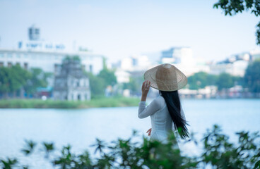 A young Vietnamese woman in a white ao dai and a conical hat standing by Hoan Kiem Lake in Hanoi, Vietnam