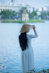 A young Vietnamese woman in a white ao dai and a conical hat standing by Hoan Kiem Lake in Hanoi, Vietnam