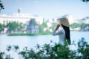 A young Vietnamese woman in a white ao dai and a conical hat standing by Hoan Kiem Lake in Hanoi, Vietnam