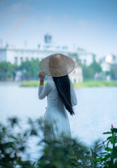 A young Vietnamese woman in a white ao dai and a conical hat standing by Hoan Kiem Lake in Hanoi, Vietnam
