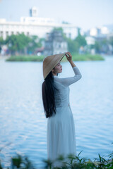 A young Vietnamese woman in a white ao dai and a conical hat standing by Hoan Kiem Lake in Hanoi, Vietnam