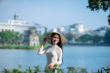 A young Vietnamese woman in a white ao dai and a conical hat standing by Hoan Kiem Lake in Hanoi, Vietnam