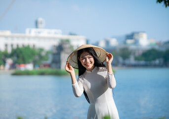 A young Vietnamese woman in a white ao dai and a conical hat standing by Hoan Kiem Lake in Hanoi, Vietnam